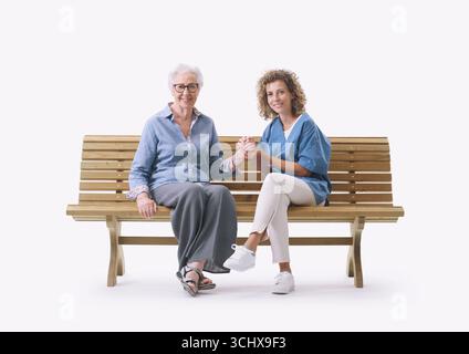 Friendly nurse sitting on a bench with a senior lady and comforting her, she is holding her hand, isolated on white background Stock Photo
