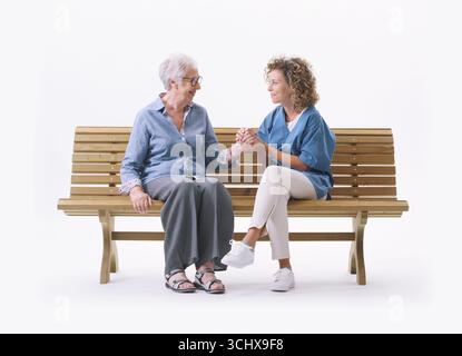Friendly nurse sitting on a bench with a senior lady and comforting her, she is holding her hand, isolated on white background Stock Photo
