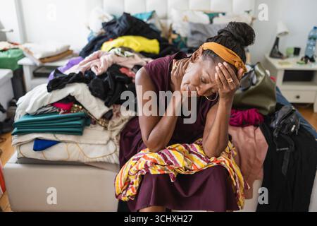 Tired woman sitting on bed with pile of clothes in bedroom Stock Photo