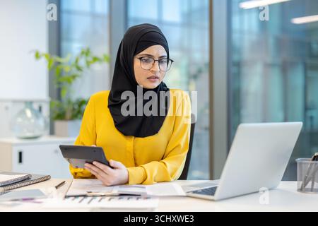 Young muslim woman wearing hijab and glasses, working as an accountant, using a calculator and laptop to manage financial data, sitting at her desk in a modern office, concentrating on her tasks Stock Photo