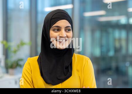 Confident and successful businesswoman wearing a hijab smiles while standing in a modern office environment, representing diversity and inclusion in the workplace Stock Photo