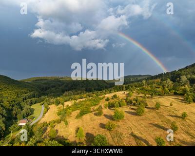 Aerial view of a vibrant rainbow arcing across a landscape of rolling hills and dark storm clouds, painting the sky with hope, Čačín, Banskobystrický kraj, Slovakia. Stock Photo