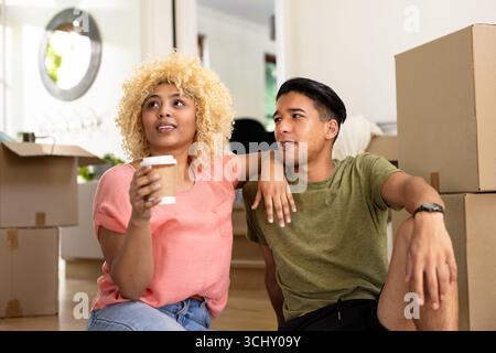 Young diverse couple enjoying coffee while unpacking boxes in new home, feeling relaxed Stock Photo