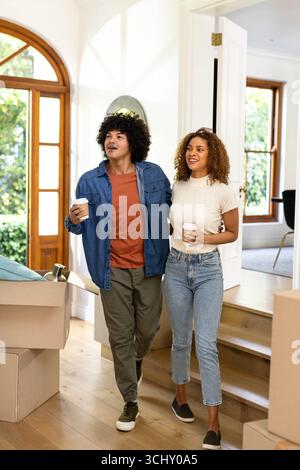 Young diverse couple carrying coffee cups, smiling while moving into new home with boxes Stock Photo