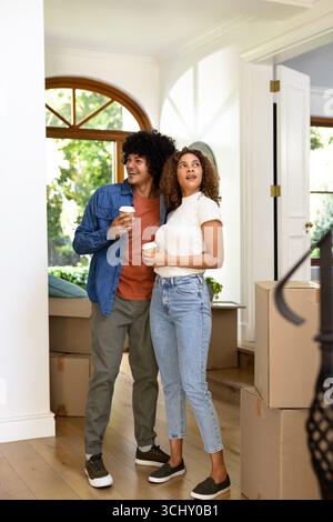 Young diverse couple holding coffee cups, standing among moving boxes at new home Stock Photo