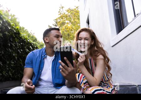Young diverse couple sitting in garden, smiling and holding pregnancy test with smartphone Stock Photo