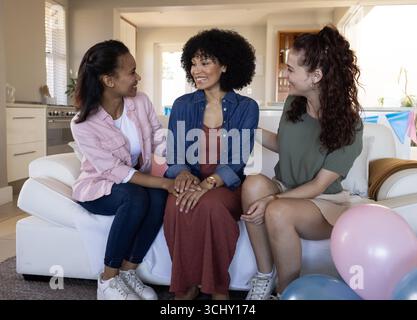 Three diverse female friends sitting on couch, smiling and celebrating friendship at home Stock Photo