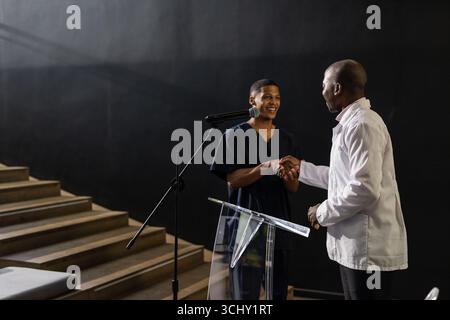 Two men shaking hands on stage, smiling and engaging in conversation Stock Photo
