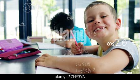 Smiling boy holding pencil and writing on notebooks and math sheets at classroom desk, copy space Stock Photo