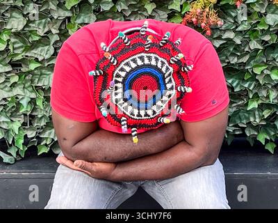 15.06.2023, Berlin, Germany, Europe - A dark-skinned man wearing a colorful knitted hat and crossed arms sits in front of a hedge in Charlottenburg. Stock Photo