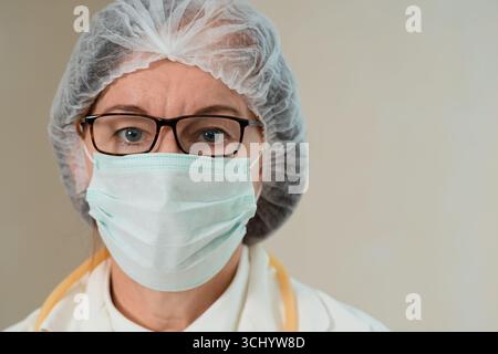 Close-up portrait of a mature female doctor wearing a medical mask and surgical cap. Her calm, focused gaze reflects professionalism, health care commitment, and patient safety. Stock Photo