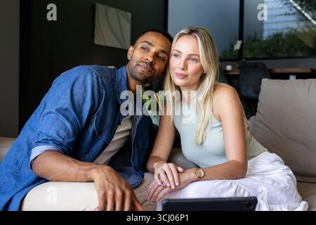 Diverse couple sitting on couch, holding hands, looking at tablet, feeling relaxed, at home Stock Photo