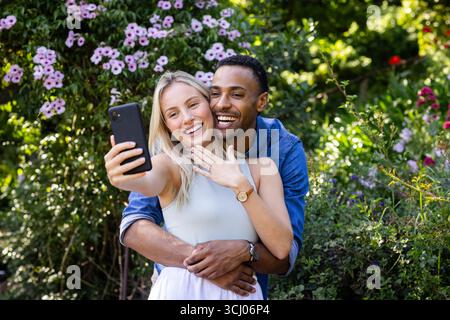 Happy diverse couple taking selfie in garden, celebrating engagement with joyful smiles Stock Photo