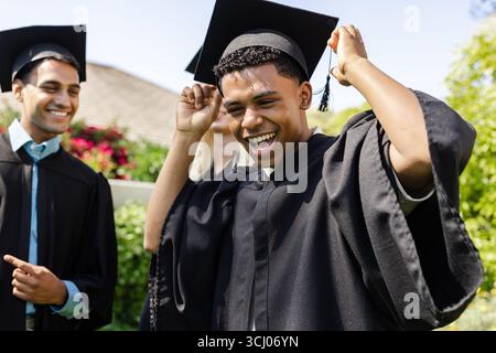 Graduating diverse students celebrating in caps and gowns, smiling joyfully in garden Stock Photo