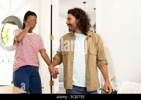 Young diverse couple holding hands, woman playfully covering eyes, entering new home Stock Photo