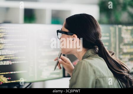 Charming female programmer analyzing code on screen in a modern workspace, focused on debugging and enhancing software Stock Photo