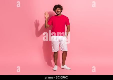 Happy young man with curly hair in red t-shirt posing on pink background while waving and smiling confidently Stock Photo