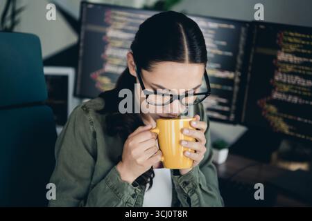 Young female programmer working remotely while enjoying coffee, with computer codes visible on multiple monitors Stock Photo