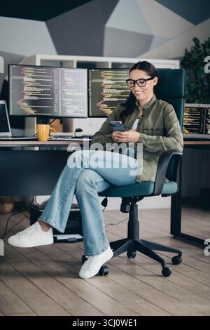 Young female programmer working on a project while using a smartphone in a modern office workspace Stock Photo