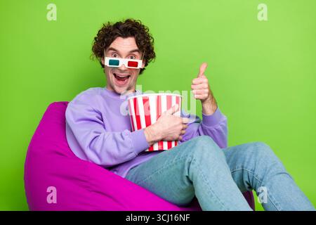 Young man in 3D glasses enjoying popcorn while seated on a bean bag showing thumbs up against green background Stock Photo