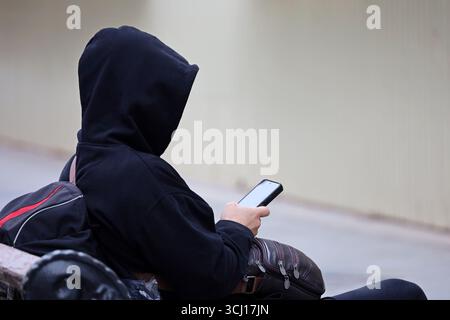 Man in hoodie using smartphone sitting on city street bench Stock Photo