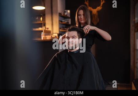 Young long haired female barber gently makes undercut hairstyle using scissors and comb bearded happy man in Salon Chair. Modern black-style barber sh Stock Photo
