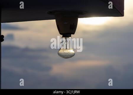 An old, dusty incandescent bulb hangs from a house roof, silhouetted against a beautiful evening sky during a vibrant sunset with warm light. Stock Photo