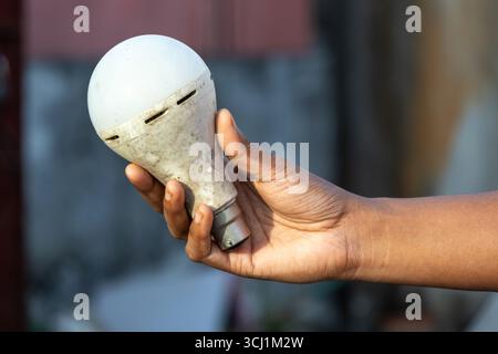 Close-up of a female hand holding a dirty, used LED light bulb against a blurred background. The old, bad bulb is a symbol of energy waste and obsoles Stock Photo