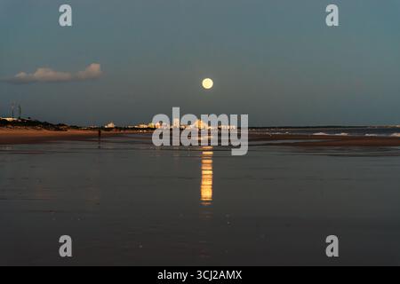 Full moon reflecting on the wet sands with a distant city skyline under a twilight sky. Stock Photo