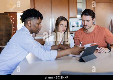 Diverse friends collaborating on project with tablet and documents at home. Collaboration, teamwork, technology, brainstorming, creativity, meeting Stock Photo