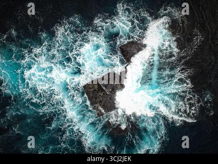 An overhead aerial view of dark, rugged rocks in the middle of a churning ocean. The powerful turquoise waves crash and swirl around the jagged outcro Stock Photo