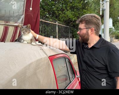 A man petting a cat resting on the roof of a red car. Human-animal connection, affection, and companionship. Stock Photo