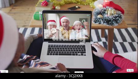 Calling father and sons wearing Santa hats holding ornaments on laptop in living room with gifts. Festive, family, holiday, warmth, connection, celebr Stock Photo