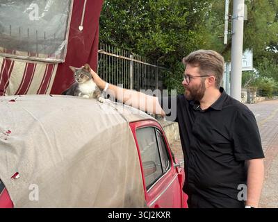 A man petting a cat resting on the roof of a red car. Human-animal connection, affection, and companionship. Stock Photo