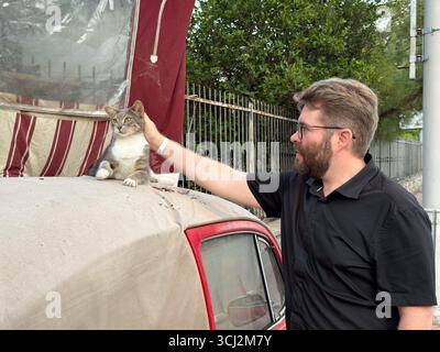A man petting a cat resting on the roof of a red car. Human-animal connection, affection, and companionship. Stock Photo