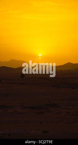Golden sunset over Moroccan desert with bright evening light, sun setting behind rolling dunes and rocky hills under warm orange sky Stock Photo