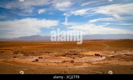 Vast arid desert plain in Morocco with scattered shrubs, dry riverbed and distant Atlas Mountains under blue sky with drifting white clouds Stock Photo