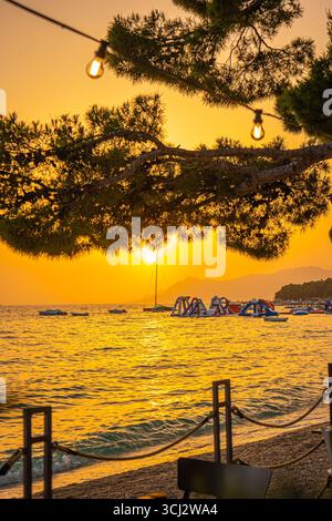 Sunset in Makarska with golden Adriatic Sea view, pine tree branches and seaside promenade with floating water park under warm orange sky Stock Photo