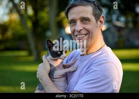 A smiling middle aged man stands in a green park holding his Canadian Sphynx cat under sunlight. The photo emphasizes joy, affection, and the bond bet Stock Photo