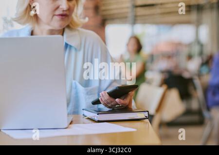 Middle Aged Woman Using Smartphone While Working on Laptop Indoors Stock Photo