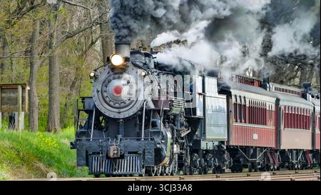 A historic steam locomotive engine pulls vintage passenger cars along tracks surrounded by lush green trees. Thick black smoke billows from the engine as it moves through the countryside. Stock Photo