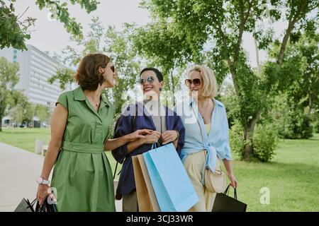 Three Middle Aged Women Chatting While Walking Outdoors with Bags Stock Photo