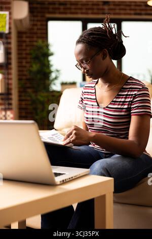 African american woman working from home, sitting on couch with clipboard in hand, reading through paperwork. Female freelancer with glasses flipping printed documents, planning and organizing tasks. Stock Photo