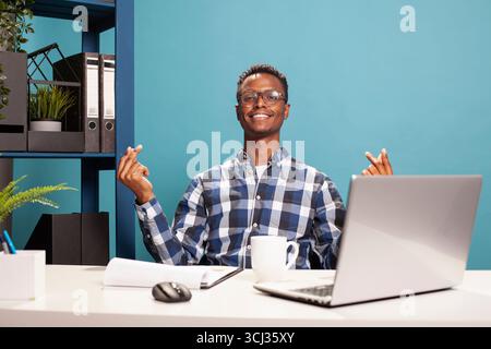 Smiling young man gesturing for cash by rubbing his fingers in a playful payday moment on a blue background. Cheerful black guy seated at his desk is expressing excitement over money rewards. Stock Photo