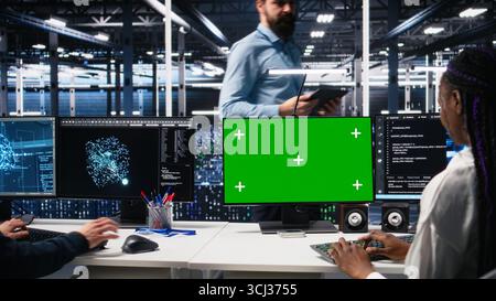 Data center engineers managing AI systems for uptime and efficiency using chroma key PC. Teamworking IT specialists using artificial intelligence on green screen computer in server hub, camera A Stock Photo
