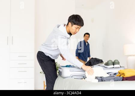 Young man packing suitcase on bed while smiling father watches in bedroom Stock Photo
