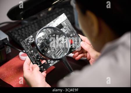 Technician inspects a circuit board using a magnifying glass during assembly in a workshop Stock Photo