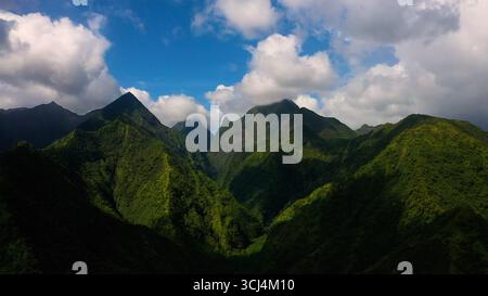 Aerial view of dramatic, lush green mountains under a bright sky dotted with fluffy white clouds, showcasing the island's rugged beauty, French Polynesia. Stock Photo