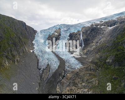 Aerial view of the icy blue glacier cascading down between the rugged, steep cliffs, a frigid spectacle against the cloudy sky, Buarbreen, Odda, Norway. Stock Photo