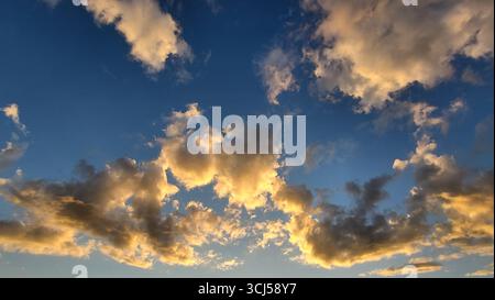 Dramatic cloud formations illuminated by warm evening sunlight during sunset, with golden-yellow tones contrasting against a deep blue sky Stock Photo
